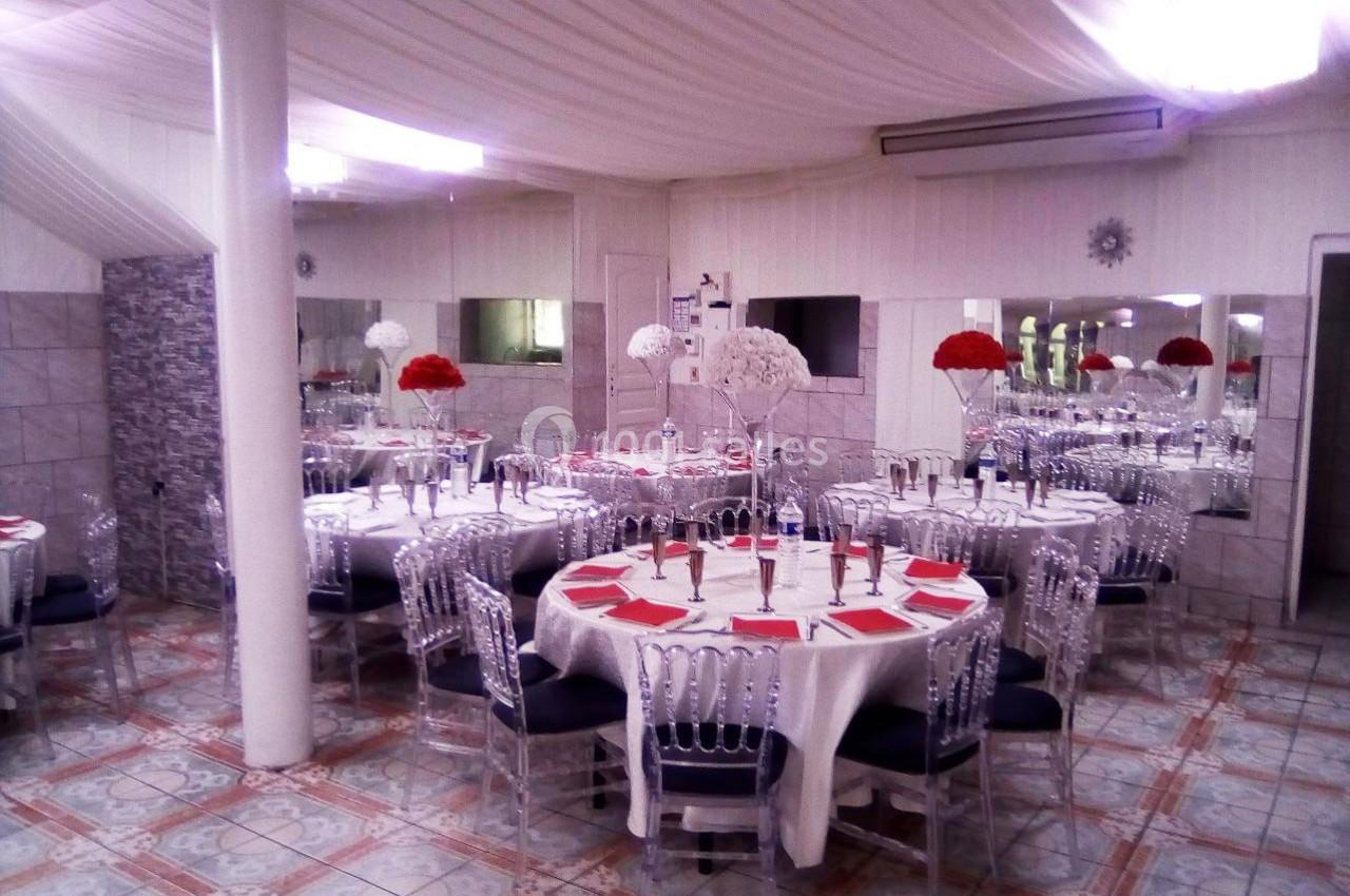 Salle de réception décorée avec des tables rondes, nappes blanches, chaises transparentes et centres de table floraux rouges…
