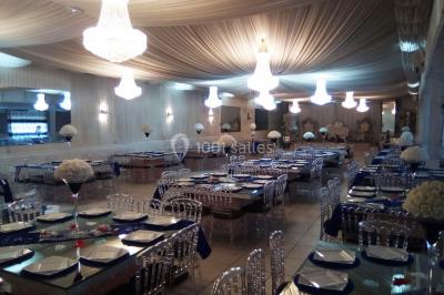 Salle de réception élégante avec tables dressées, chaises transparentes et lustres suspendus sous un plafond drapé.