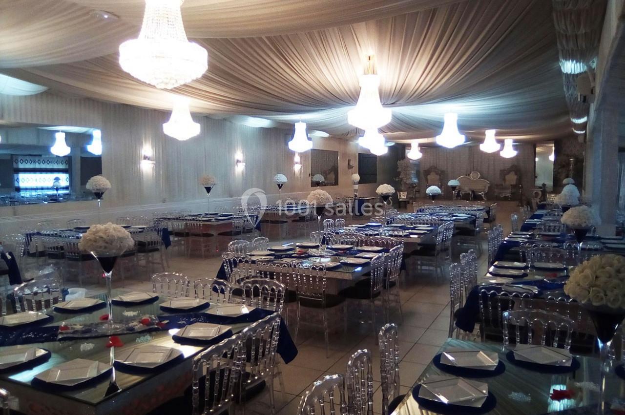 Salle de réception élégante avec tables dressées, chaises transparentes et lustres suspendus sous un plafond drapé.