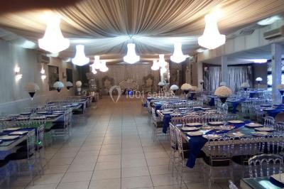 Salle de réception élégante avec tables dressées, chaises transparentes et lustres suspendus sous un plafond drapé.