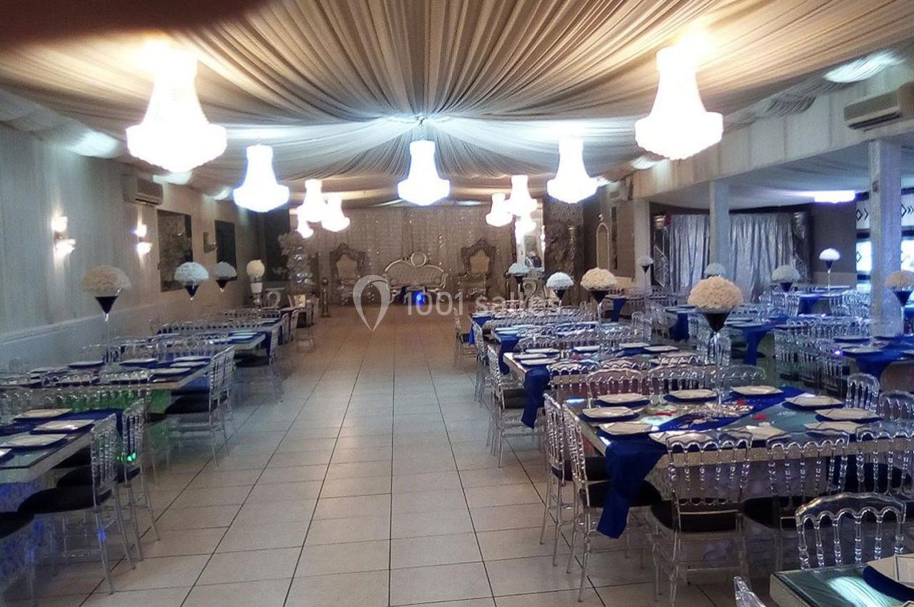 Salle de réception élégante avec tables dressées, chaises transparentes et lustres suspendus sous un plafond drapé.