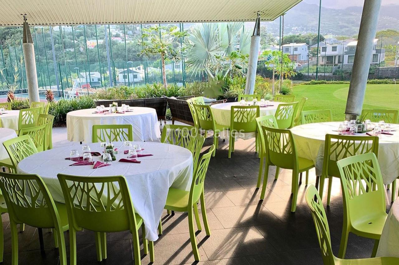 Salle à manger extérieure avec tables rondes dressées, chaises vertes et vue sur un jardin et des collines.