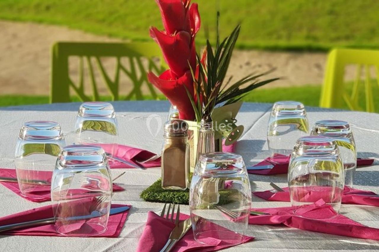 Table dressée en extérieur avec serviettes roses, verres retournés, couverts et décoration florale au centre.