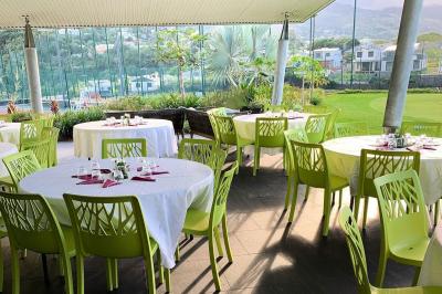 Salle à manger extérieure avec tables rondes dressées, chaises vertes et vue sur un jardin et des collines.