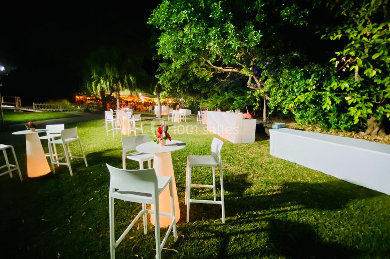 Tables hautes et chaises blanches disposées sur une pelouse éclairée, entourées de végétation et d'arbres la nuit.