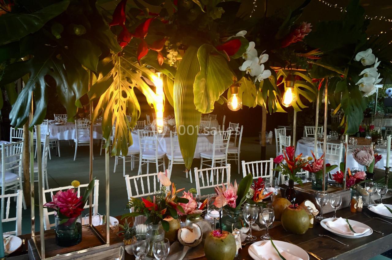 Salle de réception décorée avec des plantes tropicales, des fleurs colorées et des tables dressées élégamment.