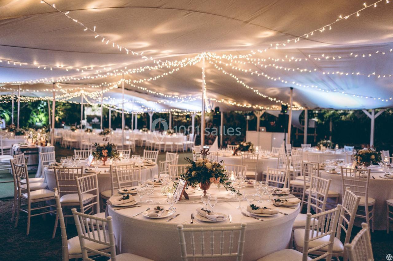Salle de réception sous une tente éclairée par des guirlandes lumineuses, avec tables rondes décorées pour un événement.