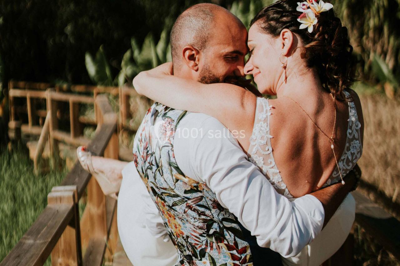 Un homme porte une femme en robe de mariée dans ses bras sur un chemin en bois entouré de végétation.
