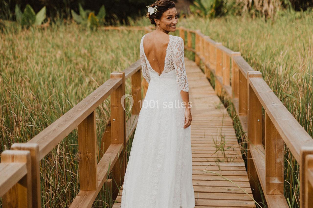 Femme en robe de mariée avec un dos ouvert, marchant sur une passerelle en bois entourée de végétation.