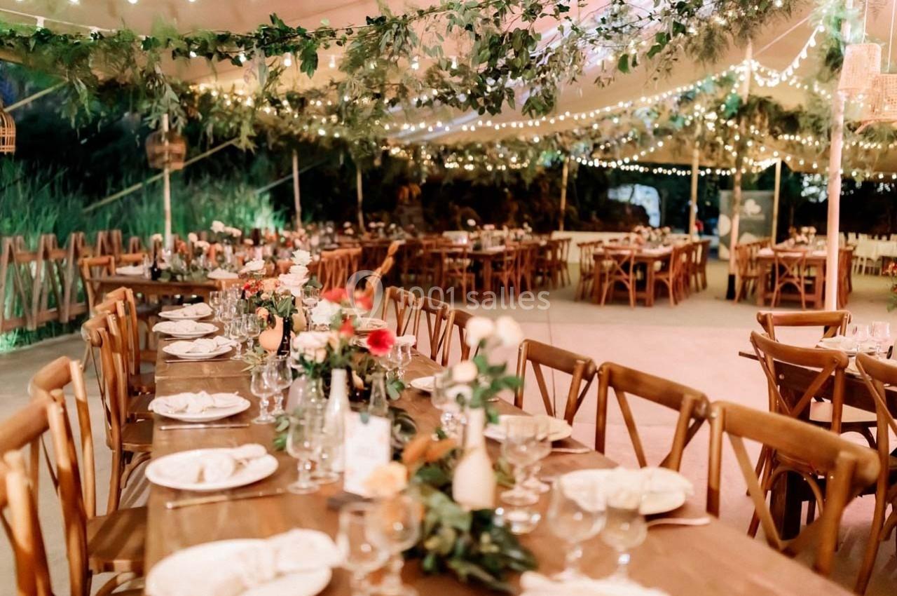 Tables en bois décorées de fleurs et de vaisselle, sous une tente éclairée par des guirlandes lumineuses.