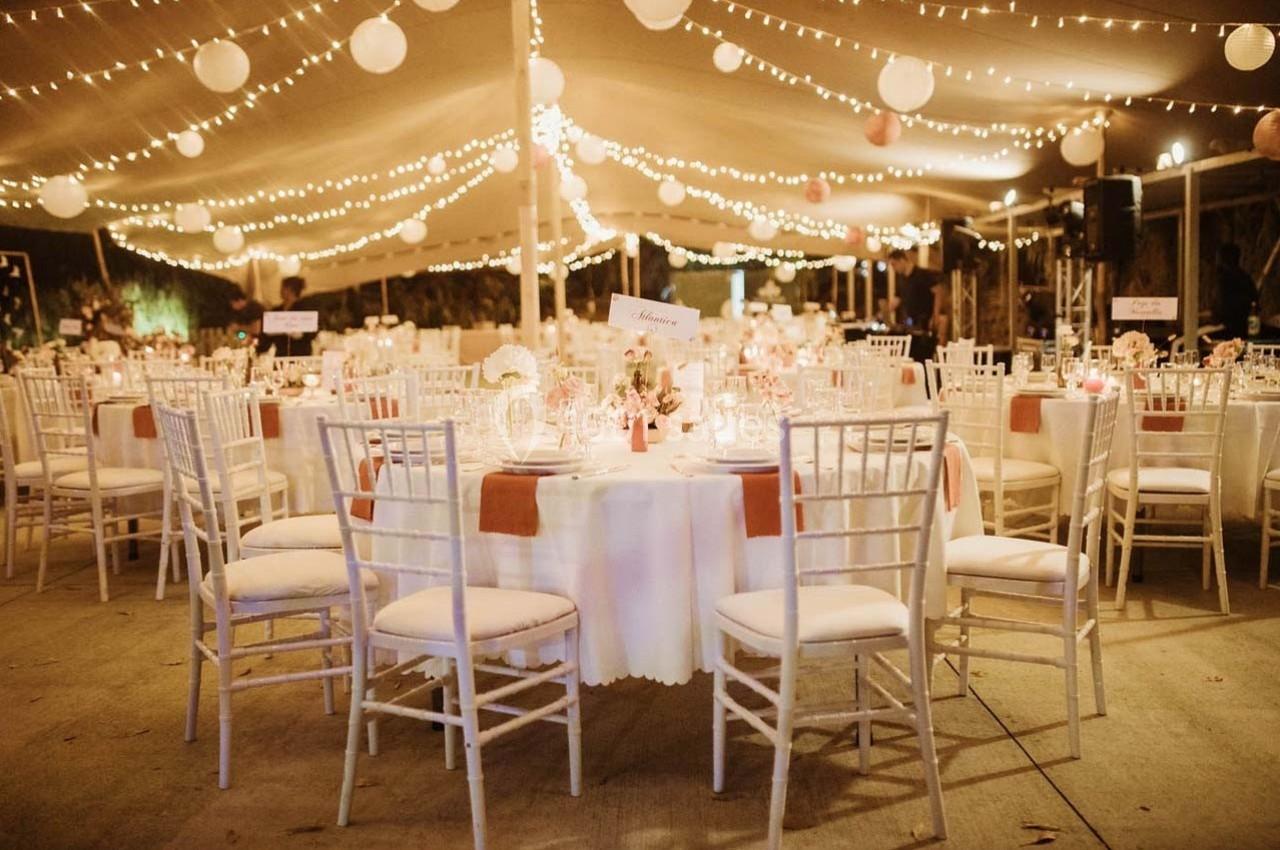 Salle de réception décorée avec des guirlandes lumineuses, tables rondes dressées et chaises blanches.