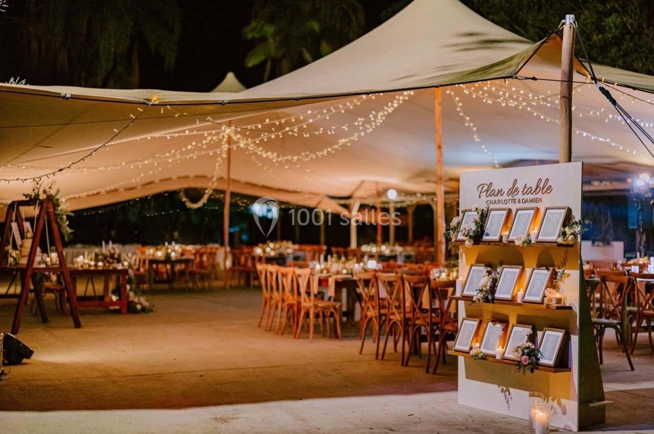 Grande tente éclairée par des guirlandes lumineuses avec des tables dressées et un plan de table décoratif au premier plan.
