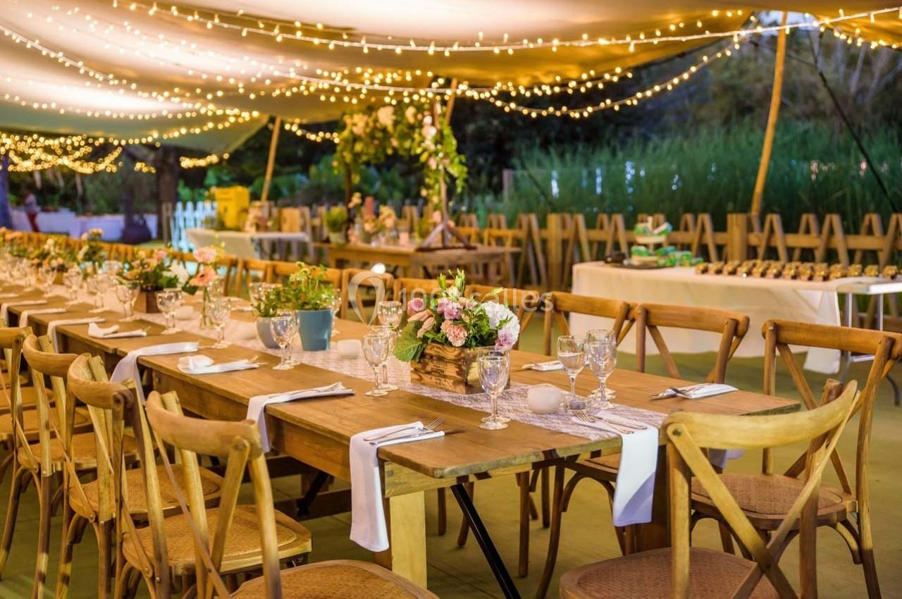 Grande table en bois décorée de fleurs et de verres sous une tente éclairée par des guirlandes lumineuses.