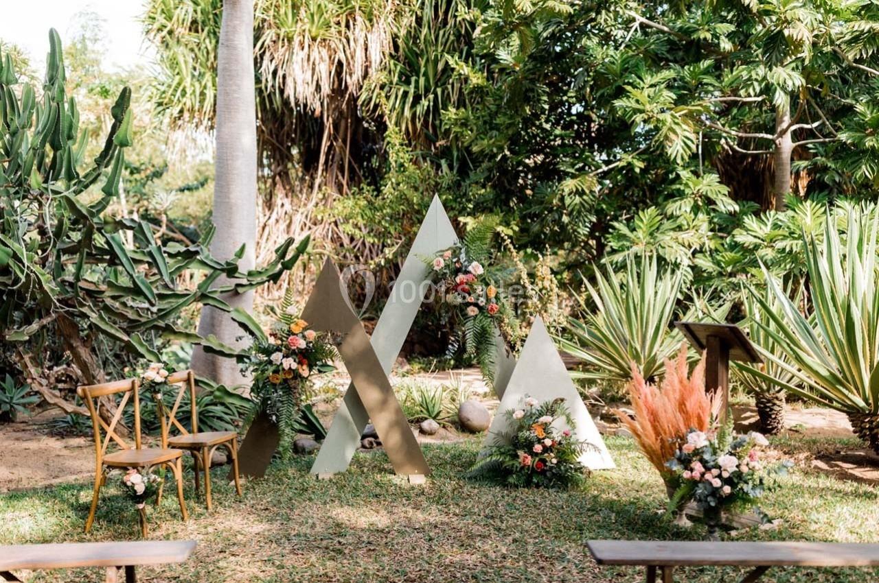 Décor de cérémonie en plein air avec chaises en bois, structures triangulaires et arrangements floraux dans un jardin…