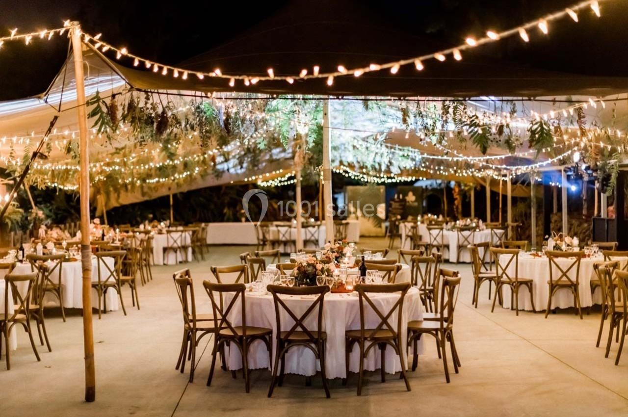 Salle de réception en plein air décorée de guirlandes lumineuses, tables rondes dressées et végétation suspendue.