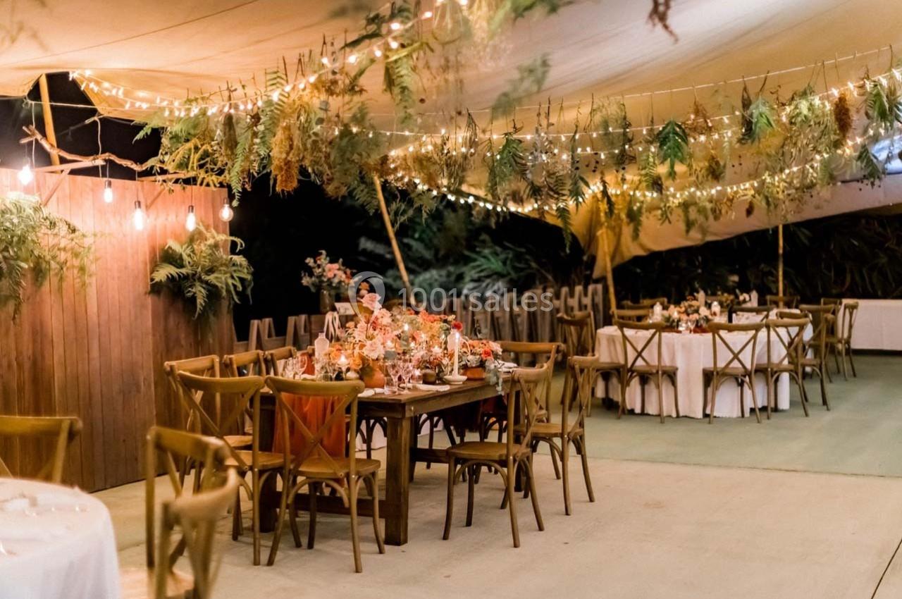 Salle de réception en extérieur décorée de guirlandes lumineuses, fleurs et feuillages suspendus, avec tables dressées.