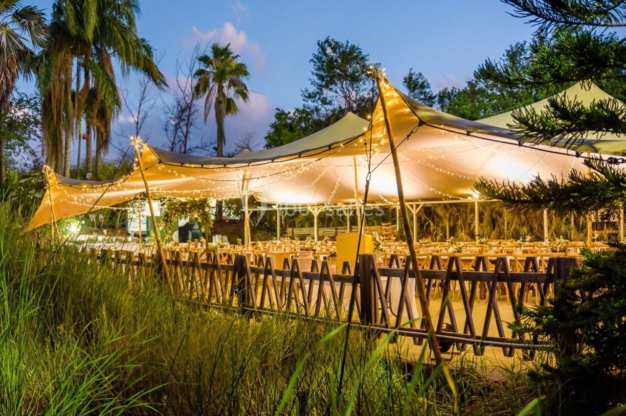 Grande tente éclairée par des guirlandes lumineuses, abritant des tables dressées dans un cadre extérieur verdoyant.