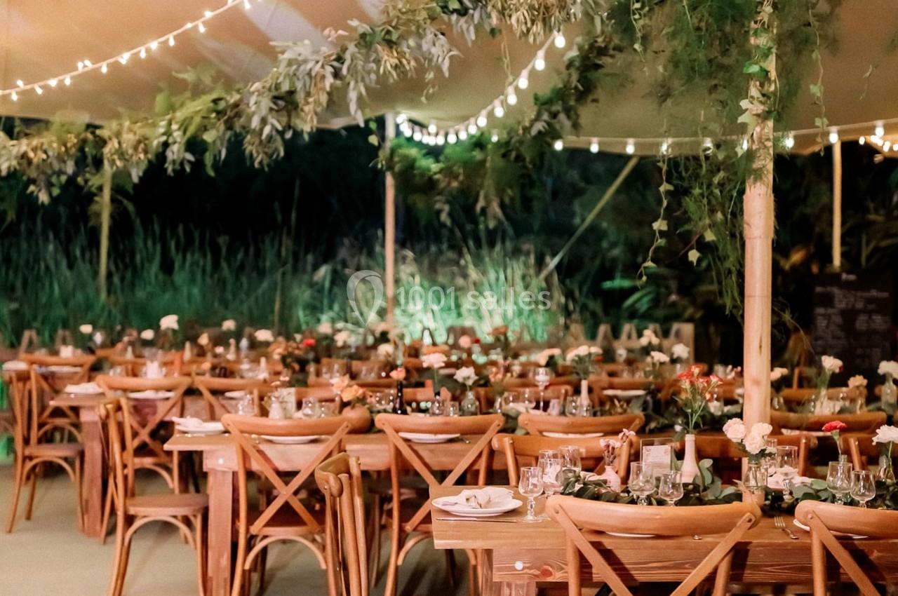 Tables en bois décorées de fleurs et vaisselle, disposées sous une tente éclairée par des guirlandes lumineuses.