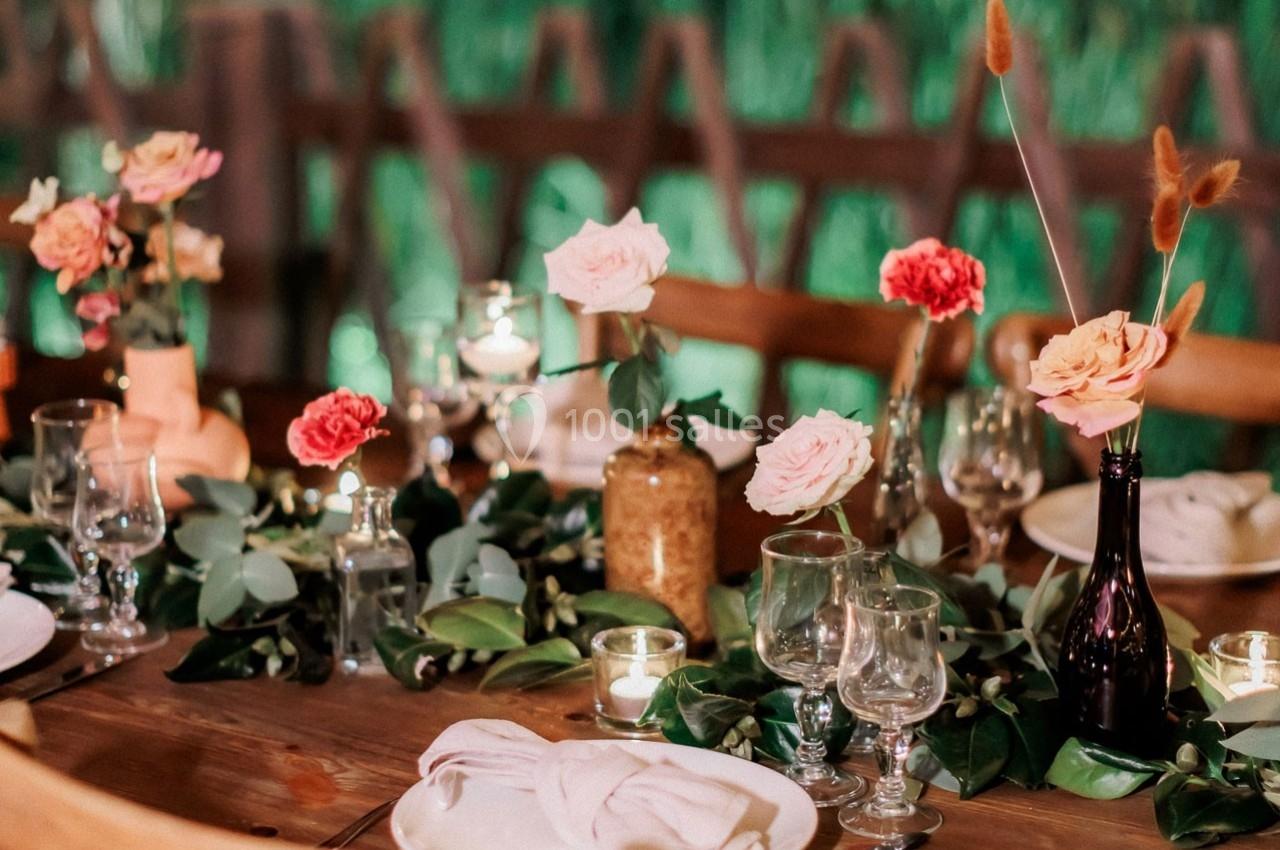 Table en bois décorée de fleurs colorées, bougies et vaisselle, avec un fond flou de verdure et de barrières en bois.