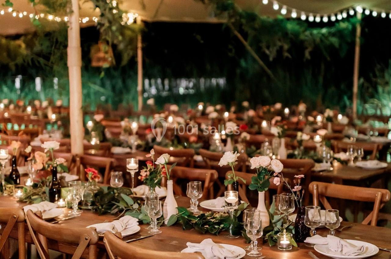 Tables en bois décorées de fleurs, bougies et vaisselle, disposées sous une tente éclairée par des guirlandes lumineuses.
