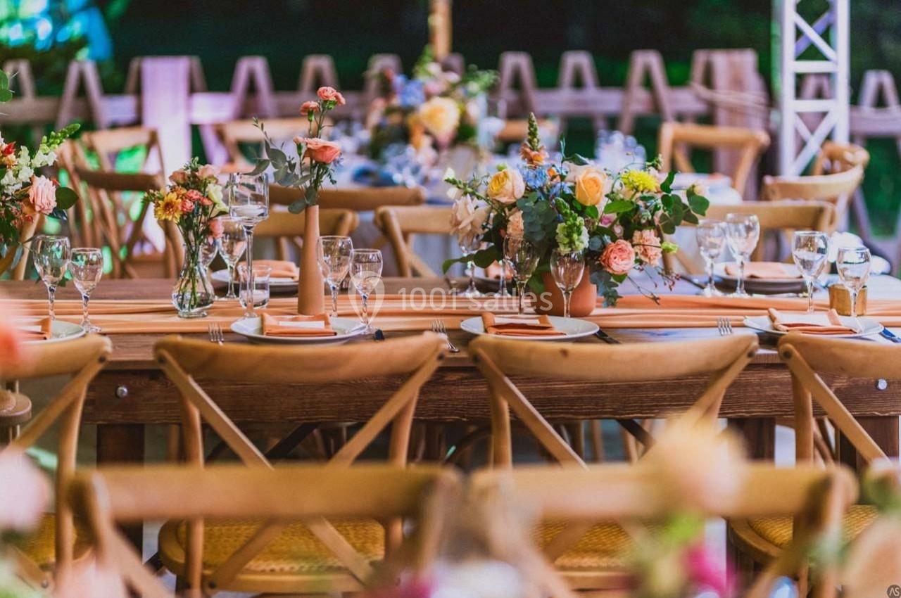 Table en bois décorée de fleurs colorées, vaisselle et verres, entourée de chaises dans un cadre extérieur.