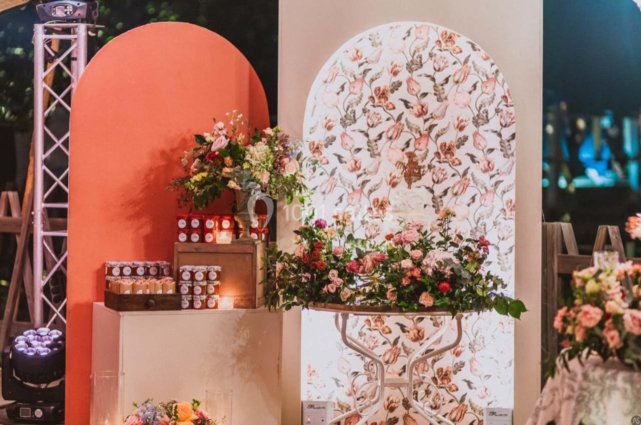 Décor floral avec table ornée de fleurs, pots de confiture alignés et panneaux colorés en arrière-plan.