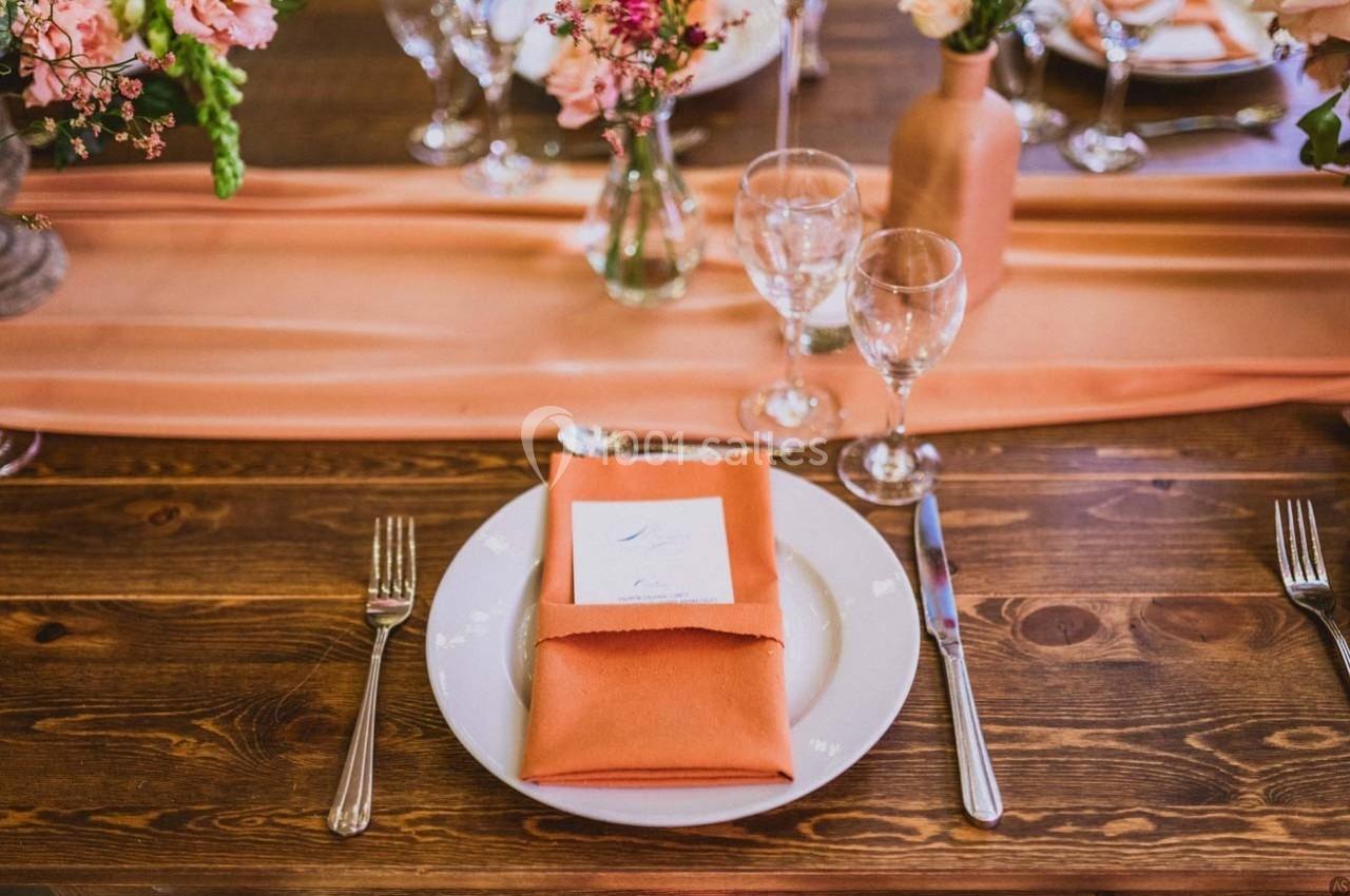Table dressée avec une assiette, une serviette orange pliée, un menu et des verres, décorée de fleurs et d'un chemin de…