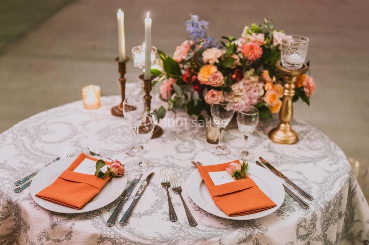 Table élégamment dressée avec nappe blanche, serviettes orange, bougies allumées et bouquet de fleurs colorées.