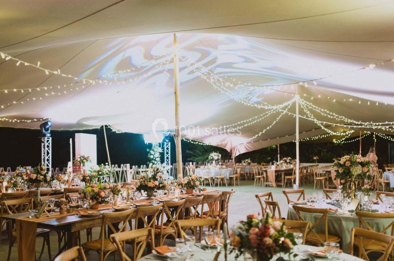 Salle de réception en plein air sous une tente illuminée, avec tables décorées de fleurs et guirlandes lumineuses.