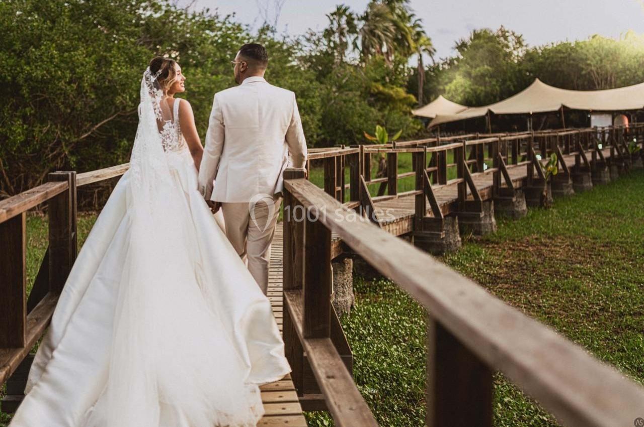 Un couple en tenue de mariage marche sur une passerelle en bois entourée de verdure et de tentes en arrière-plan.