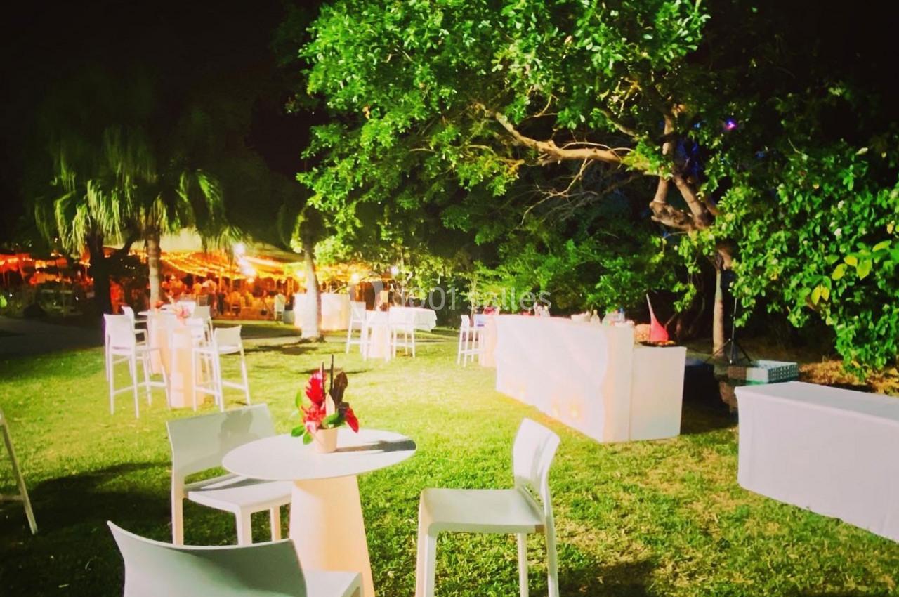 Tables et chaises blanches disposées sur une pelouse éclairée, entourées d'arbres et de lumières en arrière-plan.