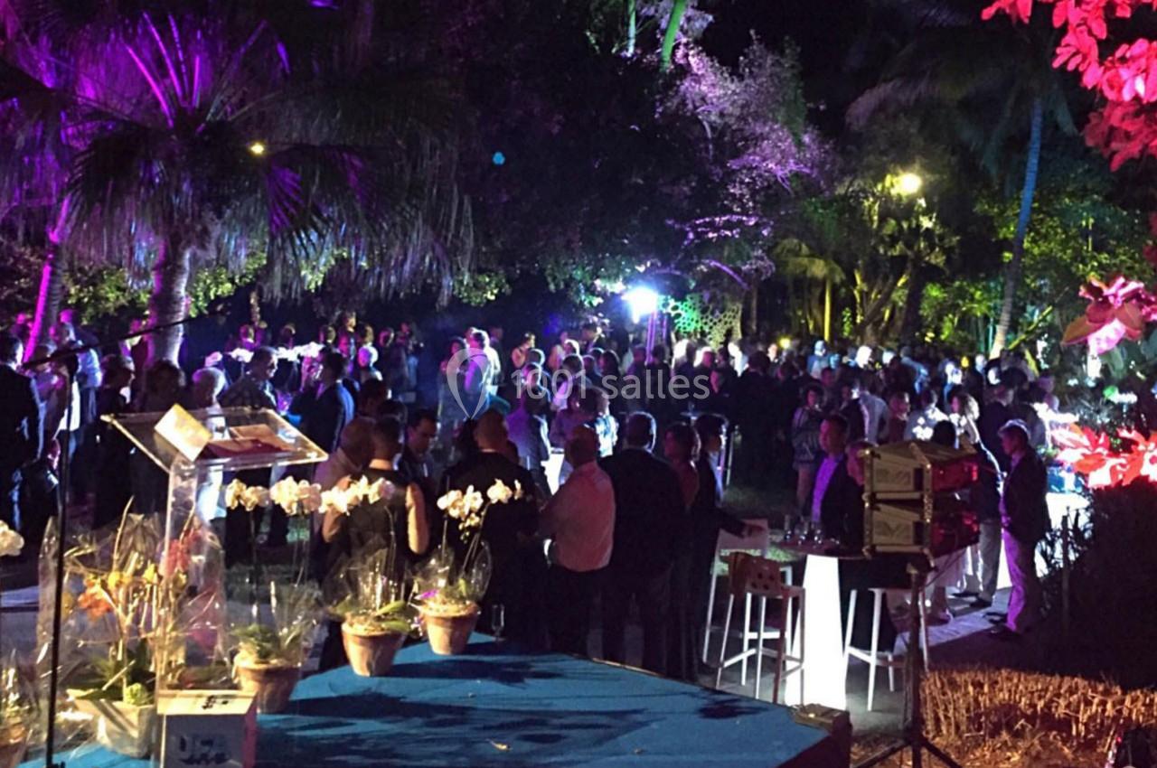 Foule rassemblée lors d'un événement nocturne en extérieur, éclairée par des lumières colorées parmi des plantes tropicales.