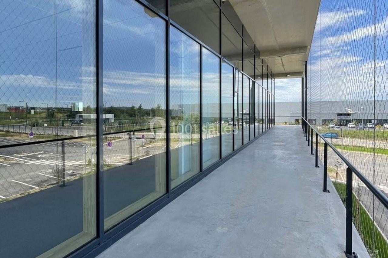 Passerelle extérieure en béton avec garde-corps, longeant une façade vitrée donnant sur un parking et un paysage dégagé.