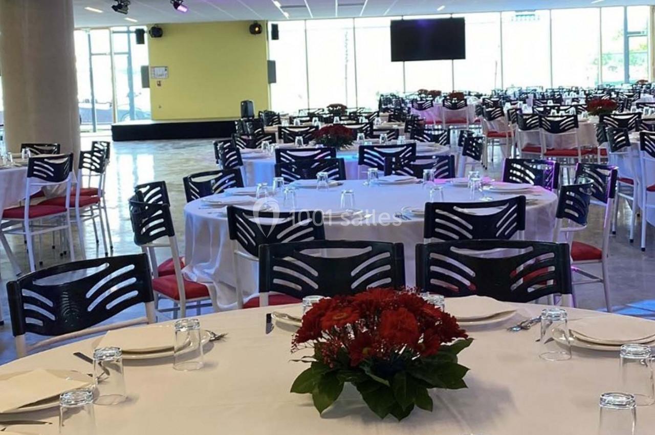 Salle de réception avec tables rondes dressées, nappes blanches, chaises noires et centres de table fleuris rouges.