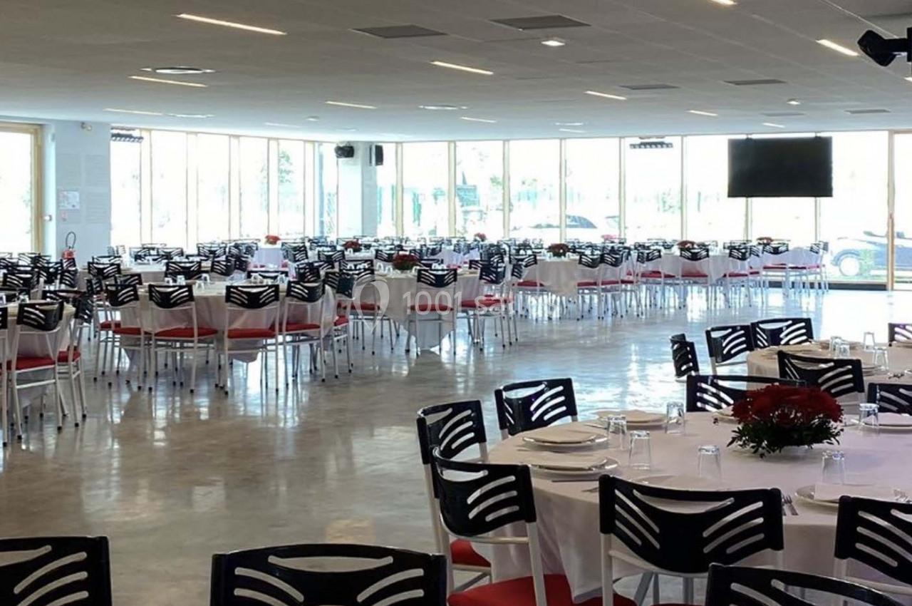 Salle lumineuse aménagée avec des tables rondes dressées pour un événement, chaises noires et rouges.