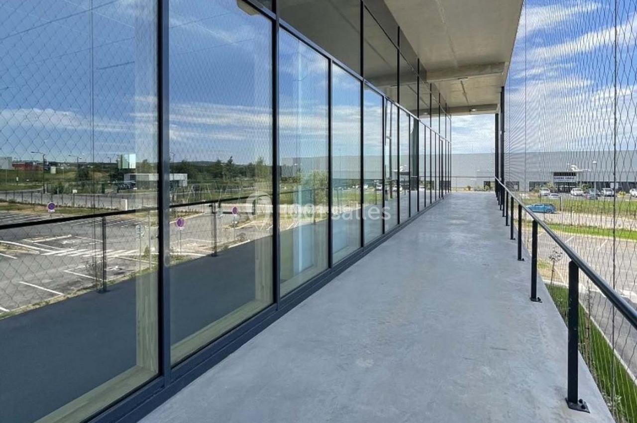 Passerelle extérieure vitrée avec vue sur un parking et un bâtiment industriel sous un ciel partiellement nuageux.