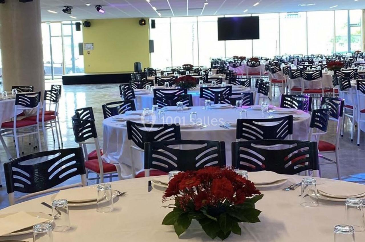 Salle de réception avec tables rondes dressées, nappes blanches, chaises noires et centres de table fleuris rouges.