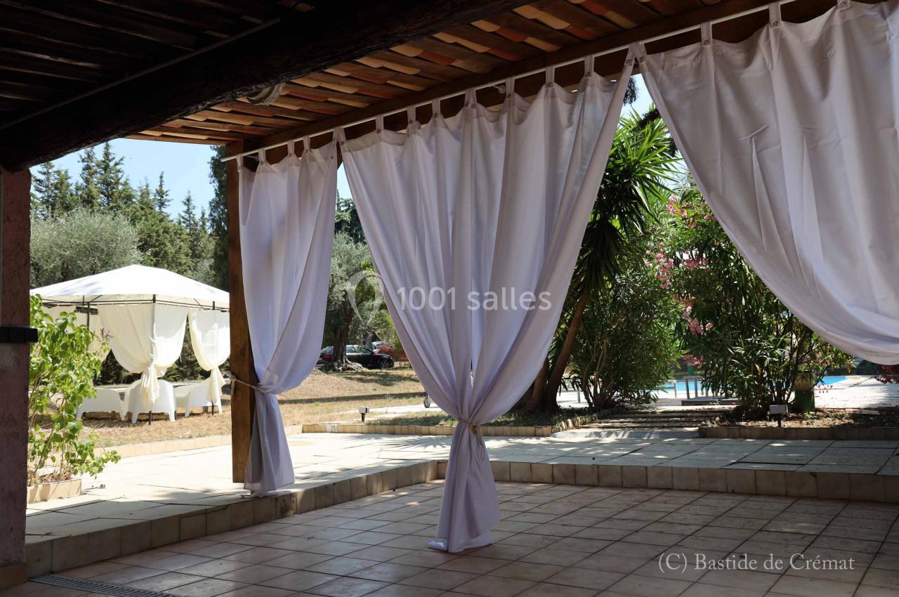 Rideaux blancs suspendus sous une pergola en bois, avec vue sur un jardin, des arbres et une piscine en arrière-plan.