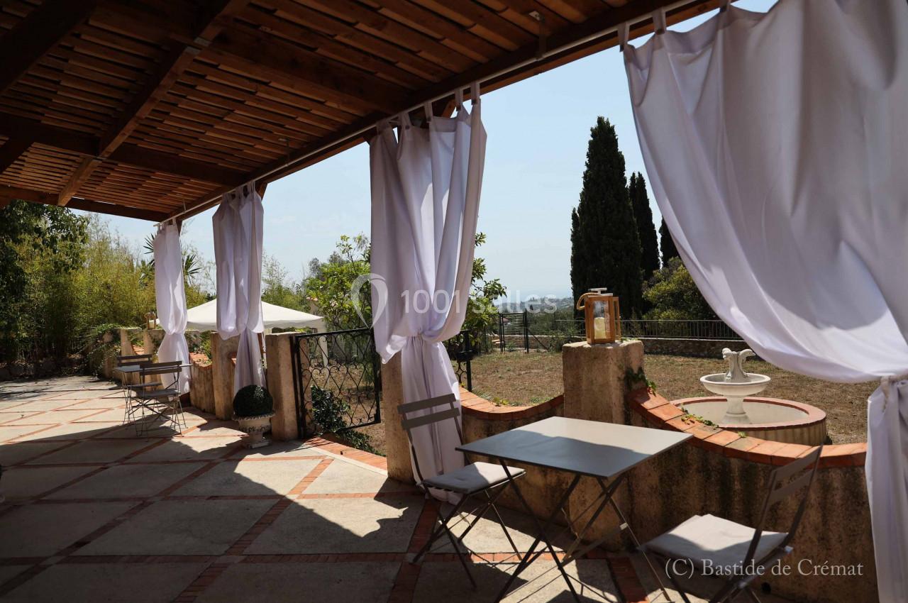 Terrasse ombragée avec rideaux blancs, tables et chaises, vue sur un jardin et paysage lointain.
