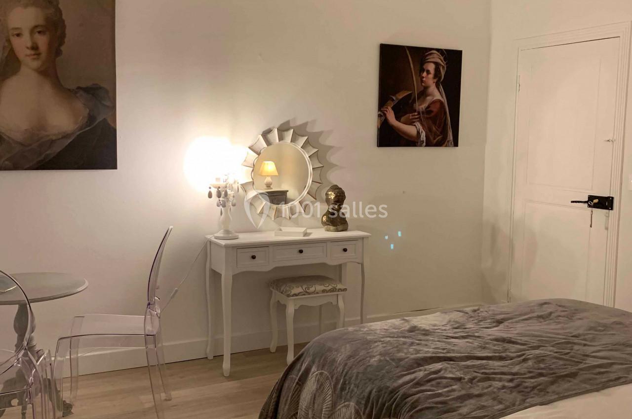 Chambre lumineuse avec un lit double, bureau blanc, chaises transparentes, tableaux et lustre décoratif.