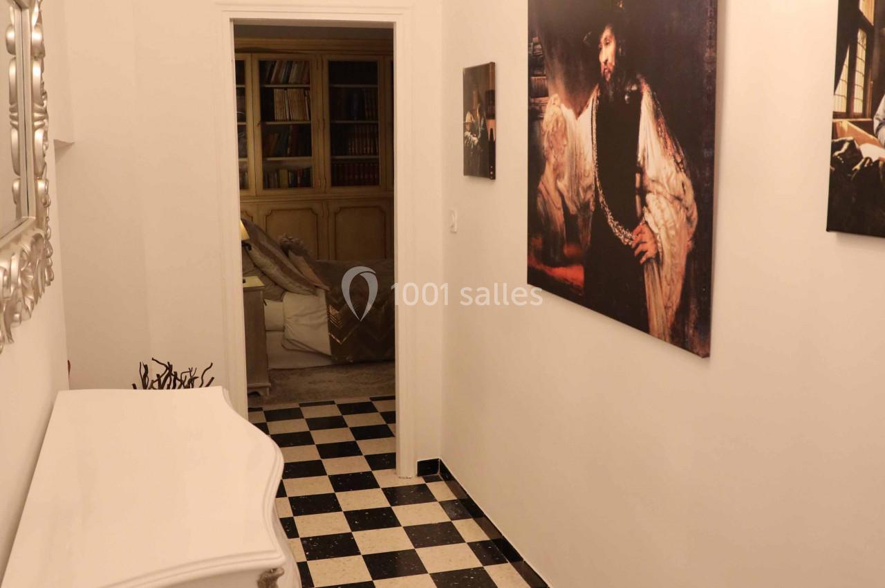 Couloir étroit avec sol en damier noir et blanc, commode blanche, lustre noir et tableaux accrochés aux murs.