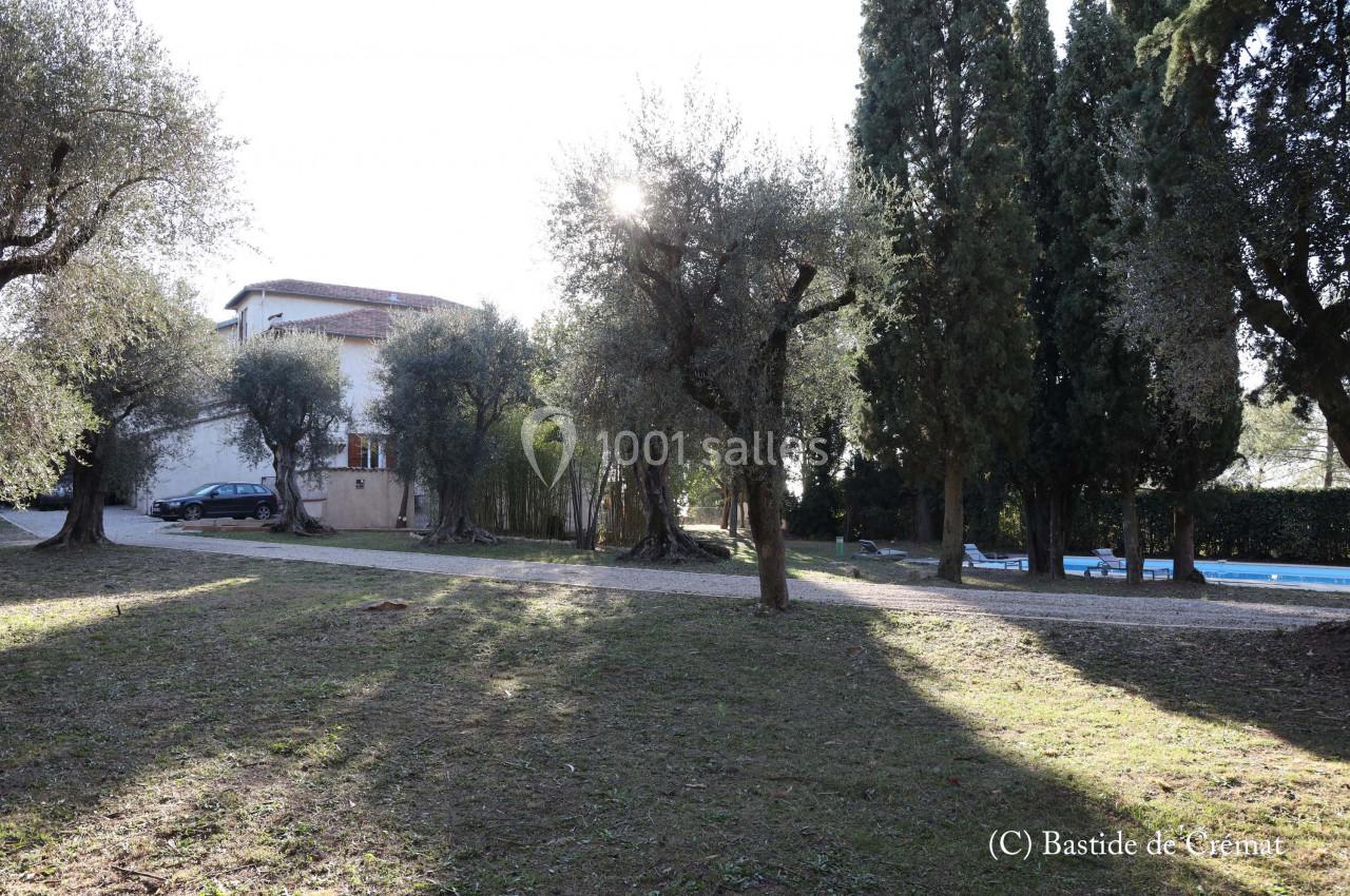 Jardin ombragé avec des oliviers, une maison blanche en arrière-plan et une piscine visible sur la droite.