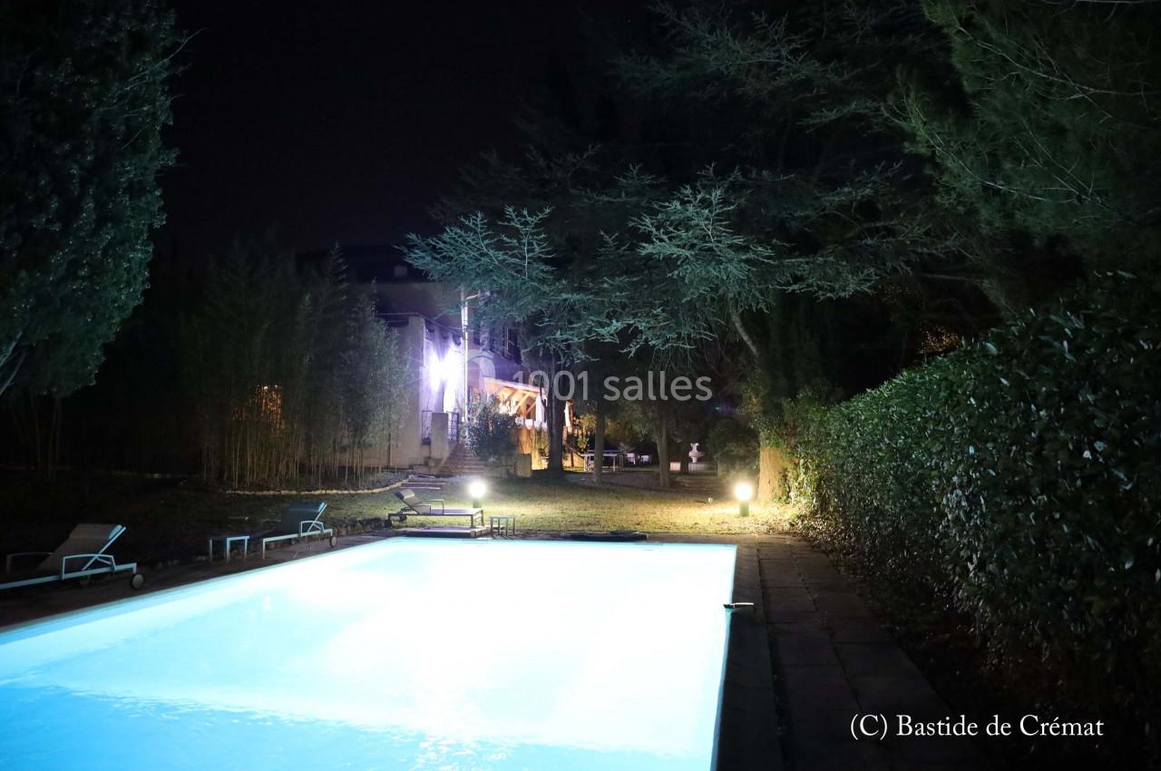 Piscine éclairée la nuit entourée de végétation et d'arbres, avec une maison visible en arrière-plan.