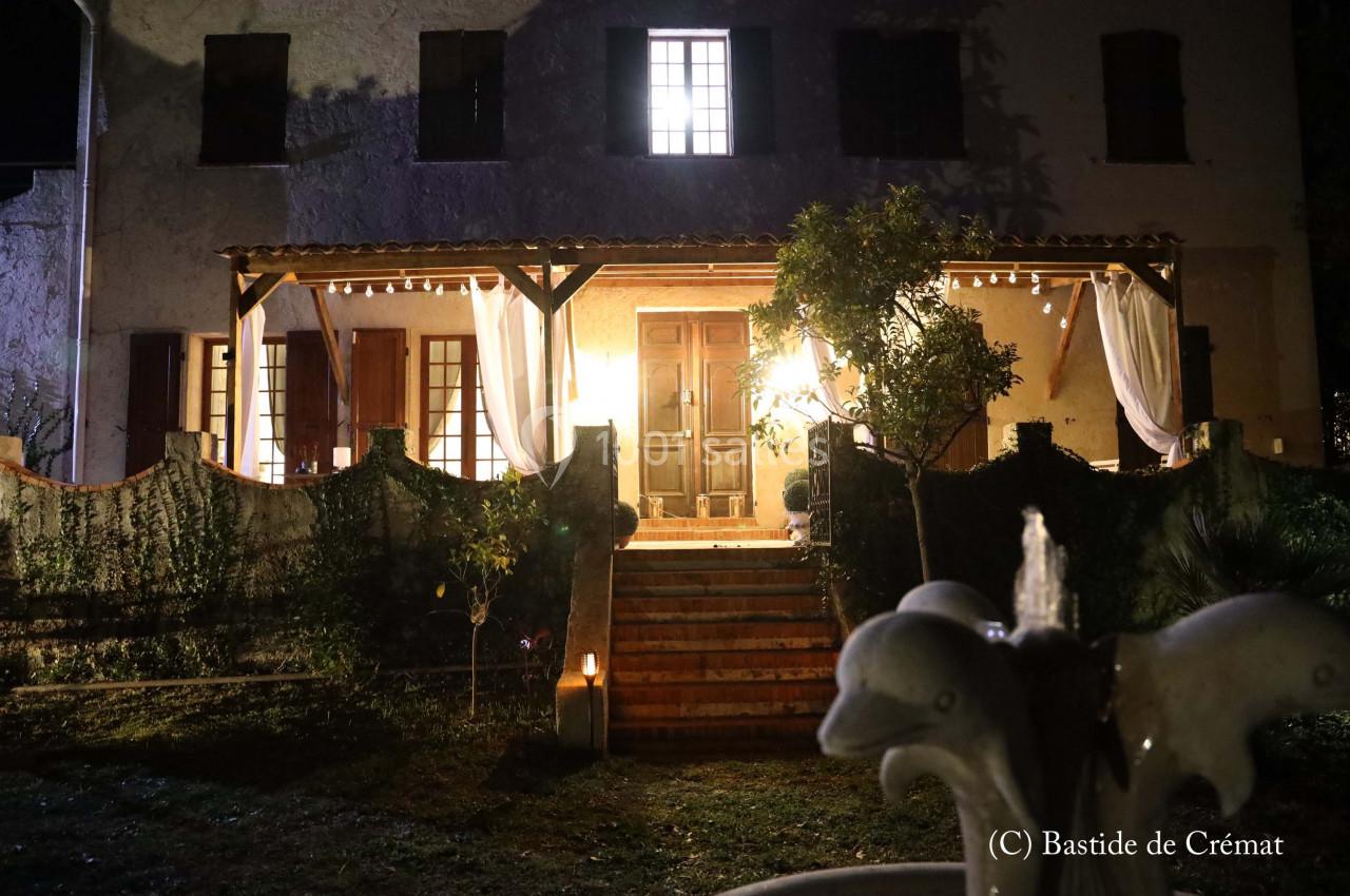 Façade d'une maison éclairée la nuit, avec terrasse illuminée, rideaux blancs et escalier en pierre.
