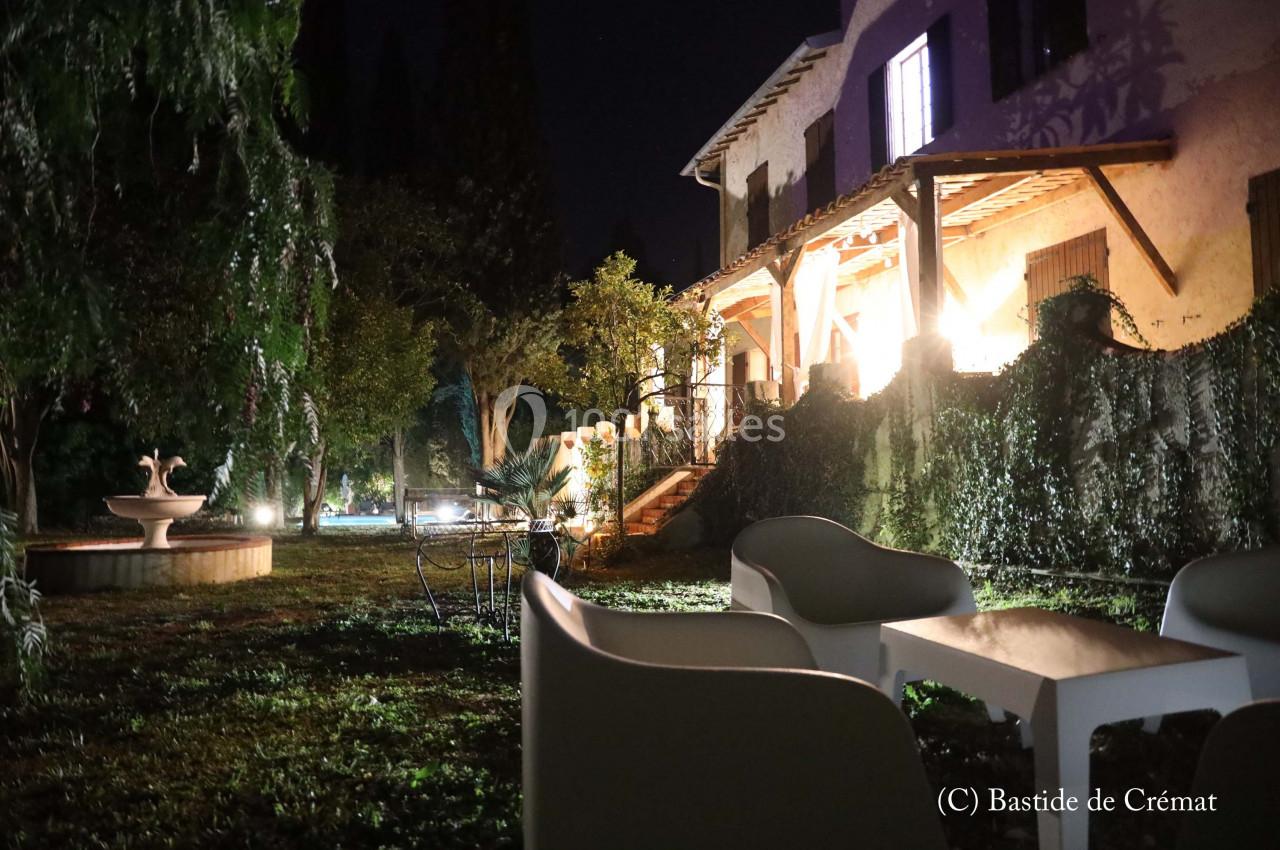 Jardin éclairé de nuit avec une fontaine, des chaises blanches et une maison aux fenêtres illuminées en arrière-plan.