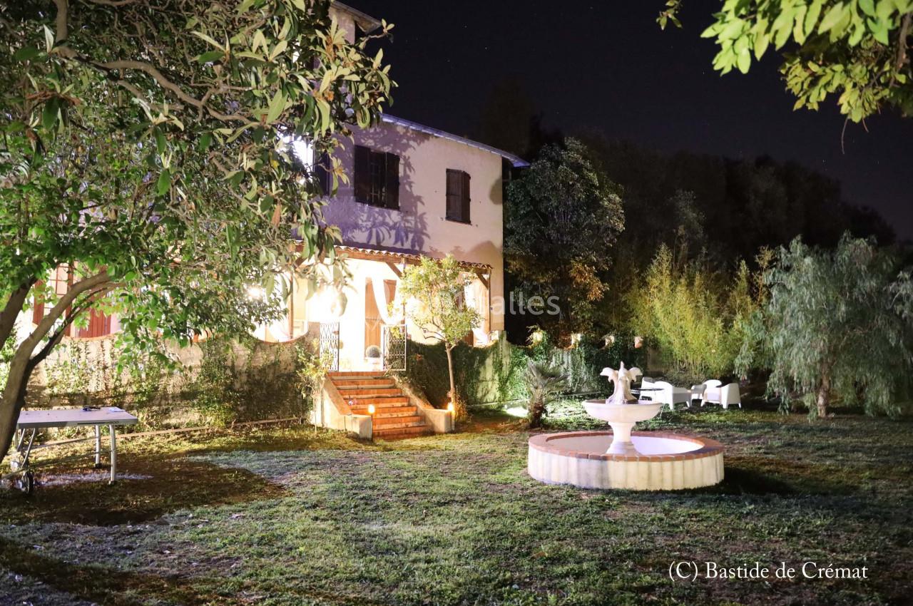 Jardin éclairé de nuit avec une fontaine centrale, des arbres et une maison en arrière-plan.