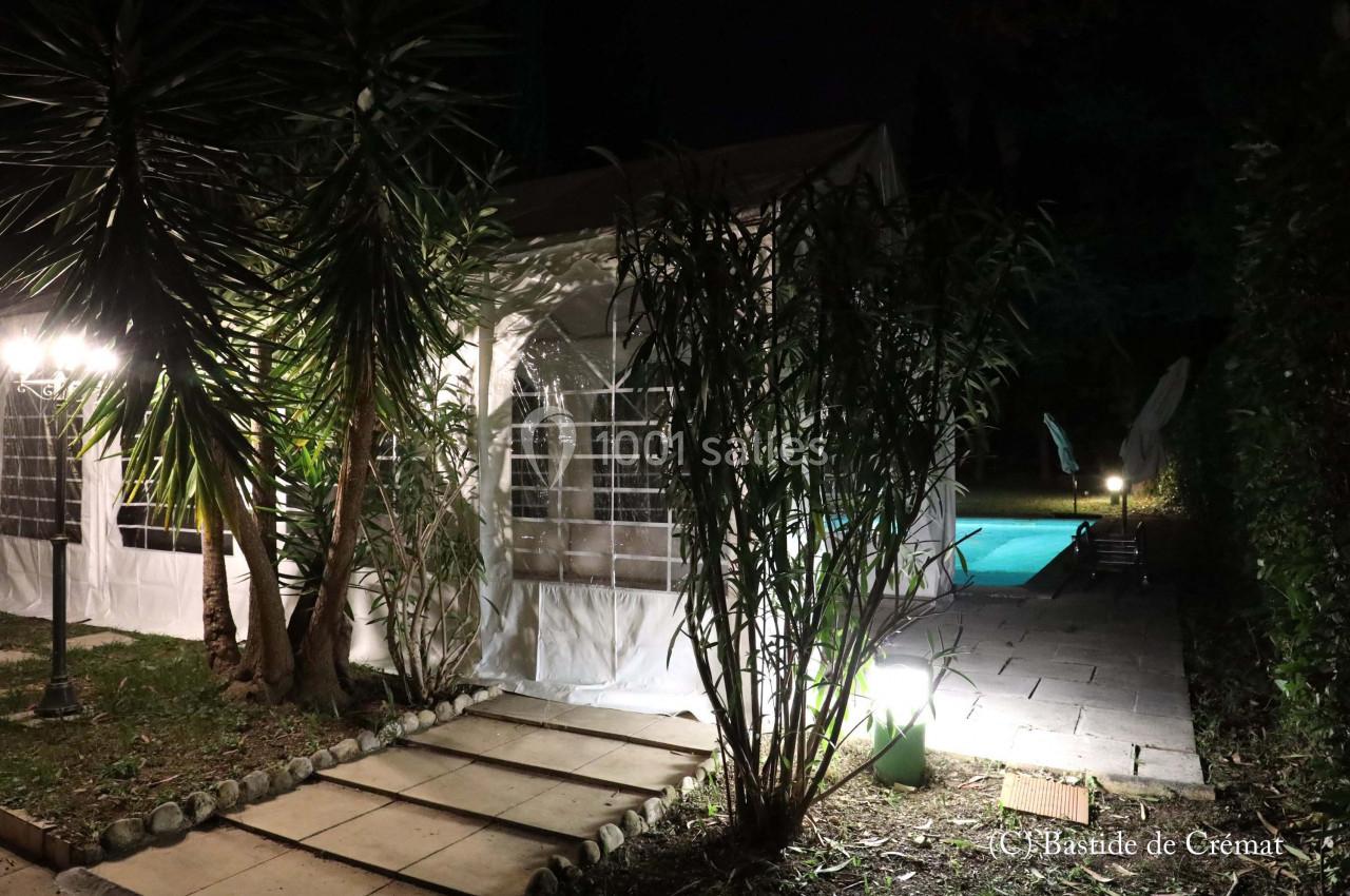 Tente éclairée de nuit près d'une piscine entourée de végétation et d'un chemin en dalles.