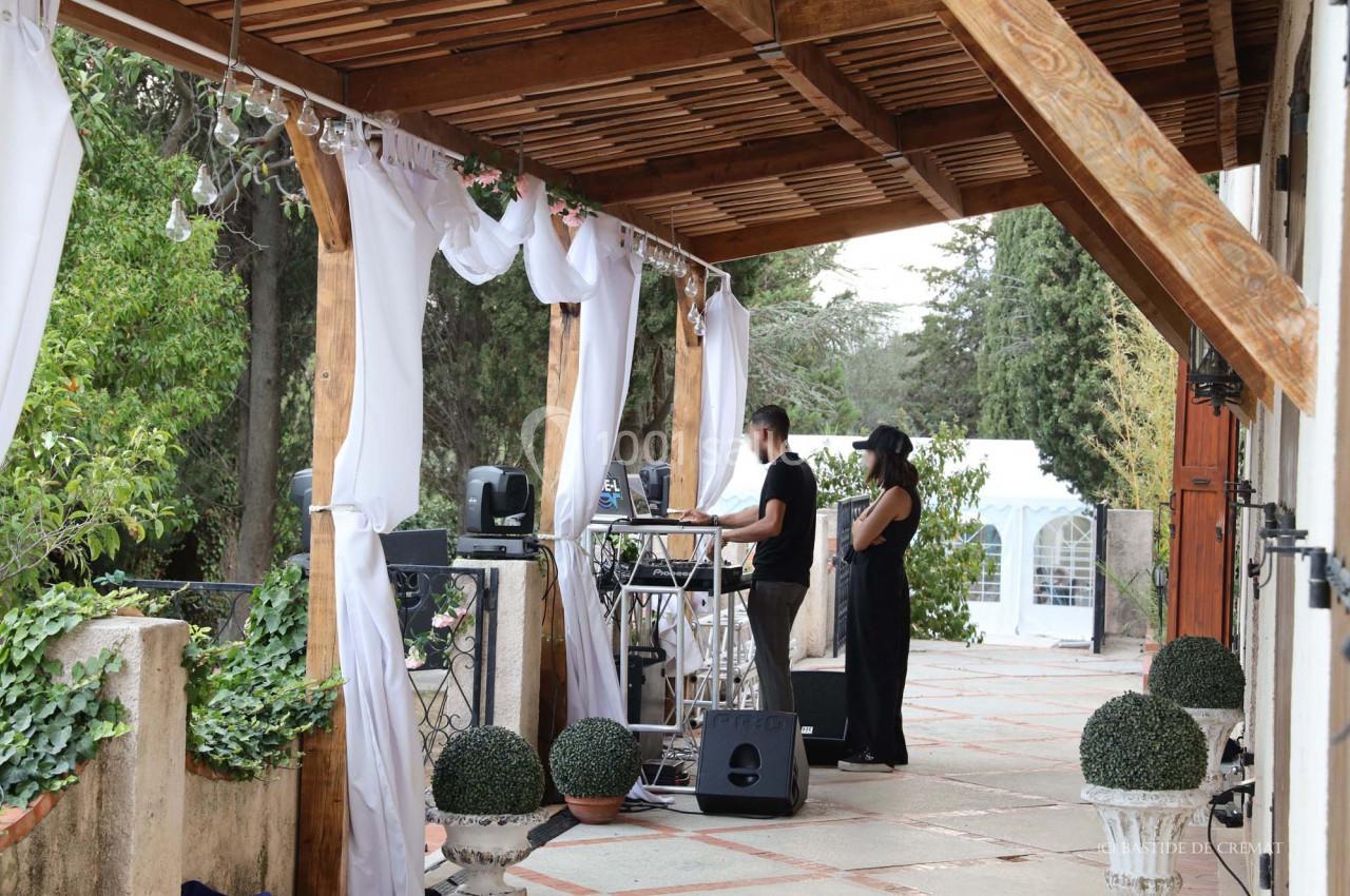 Deux personnes gèrent une table de mixage sous une pergola décorée de voilages blancs, dans un cadre extérieur verdoyant.