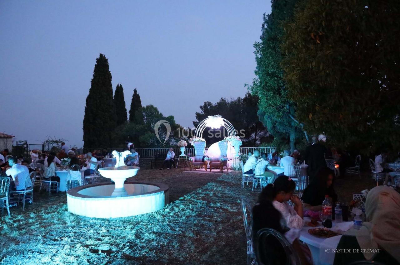 Des invités dînent en plein air dans un jardin éclairé, avec une fontaine illuminée et des arbres en arrière-plan.