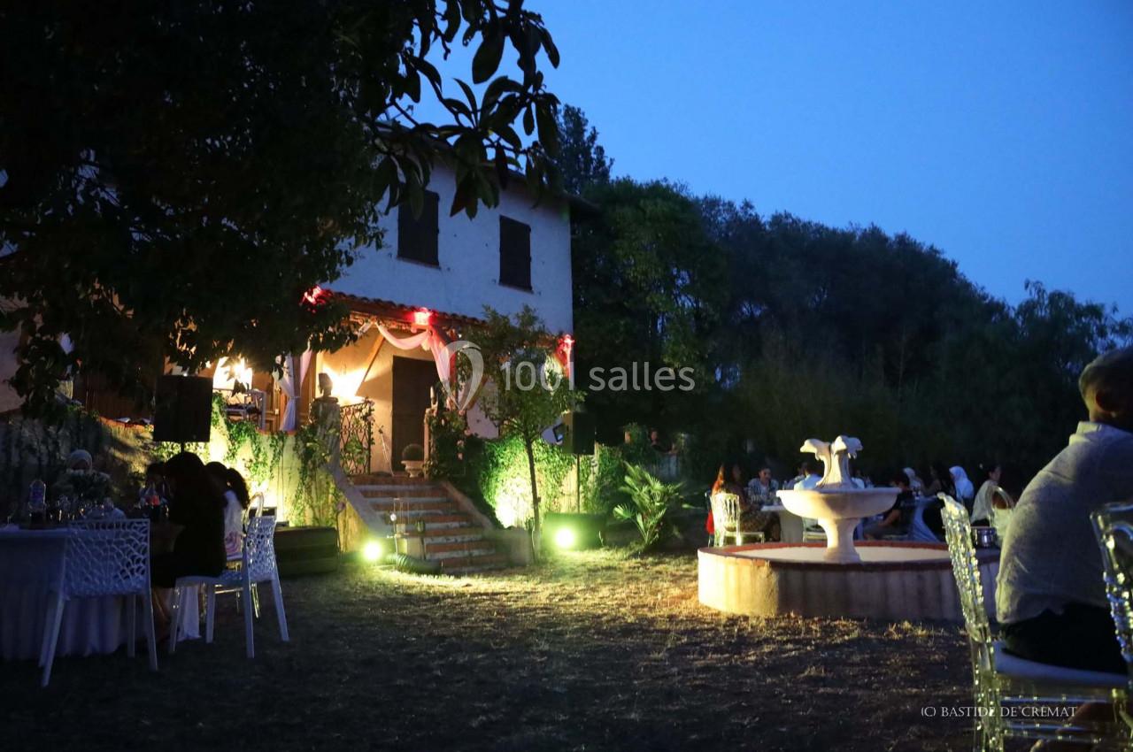 Maison éclairée en soirée avec des invités assis à des tables dans un jardin, près d'une fontaine illuminée.
